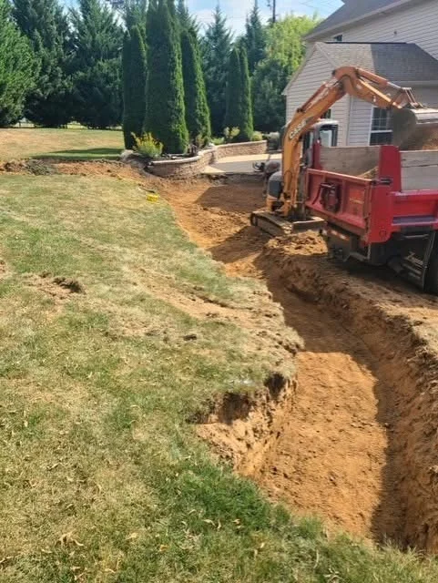 Mini excavator and dump truck digging a utility trench across a lawn, ahead of commercial property restoration